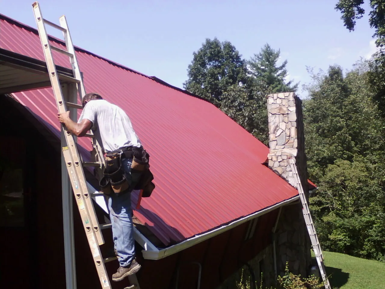 Roofer on a ladder beside a red metal roof and stone chimney — Soffit & Fascia Repair by Roofing Brothers in Hathorne